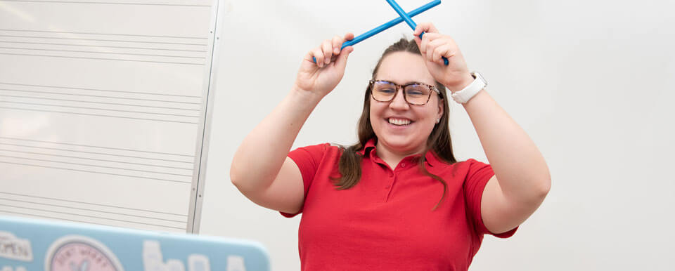 A graduate music therapy student hits drumsticks together while on a video call with a client.