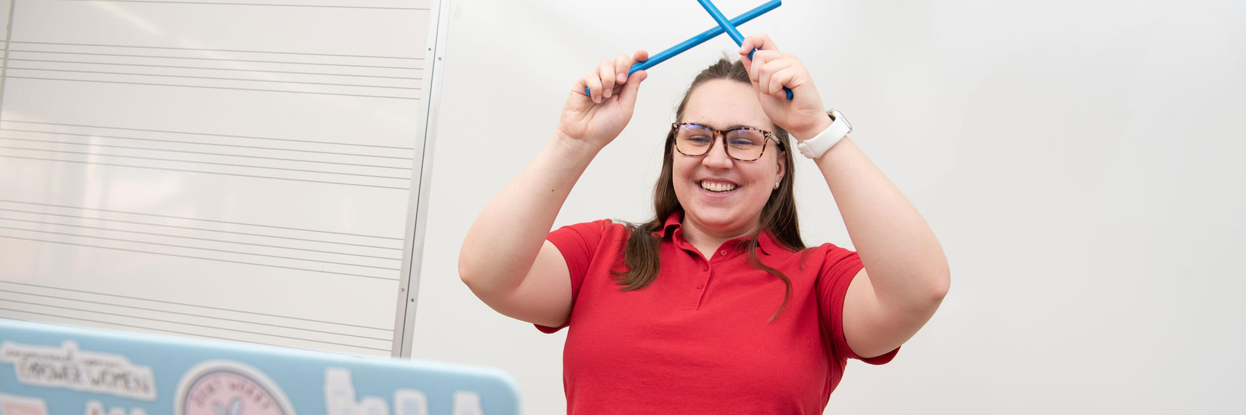 A graduate music therapy student hits drumsticks together while on a video call with a client.