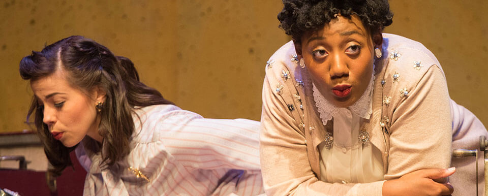 Two actresses rest their elbows on a table onstage during a performance.