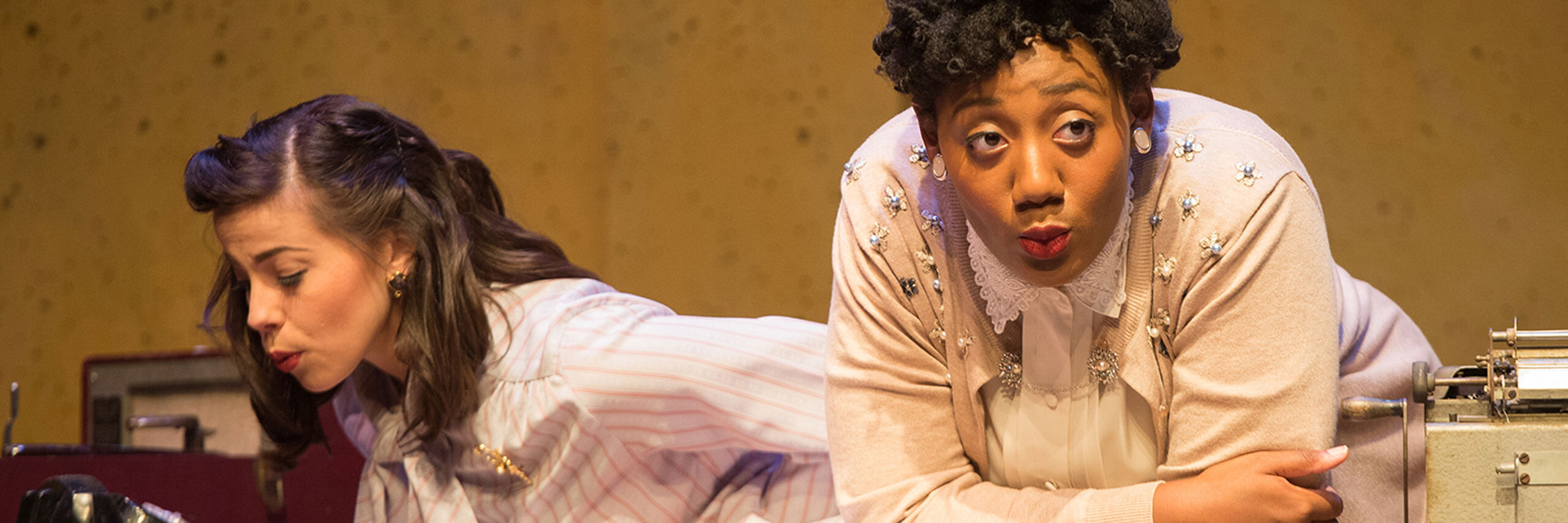 Two actresses rest their elbows on a table onstage during a performance.