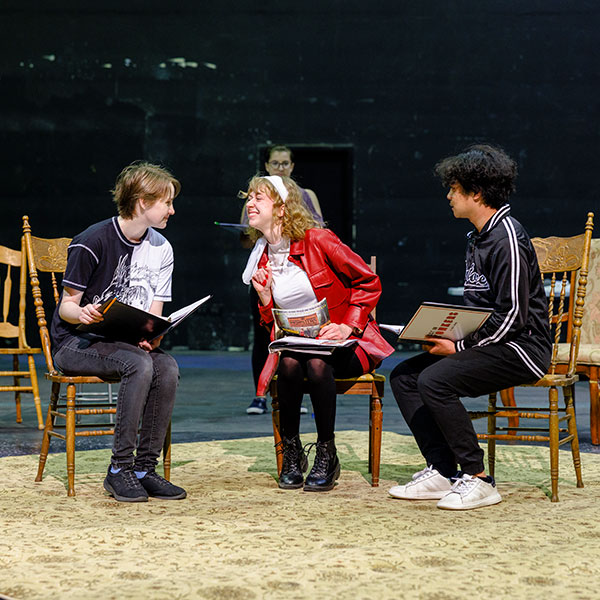 Three people sitting in chairs on a stage, holding notebooks, with a fourth person standing in the background.